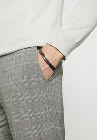 Lichtgrijze trui met ribbels bij de manchetten, gecombineerd met een geruite broek in grijs, zwart en goud, met een slim fit enzijzakken. Zwarte geweven armband.