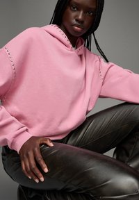 Haut à capuche rose avec des clous argentés le long des épaules, associé à un pantalon en faux cuir noir. Textures lisses, coupe décontractée et manches longues.