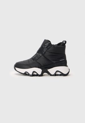 Bottine noire imperméable avec tige matelassée, semelle blanche épaisse avec motif ondulé noir et fermeture éclair latérale sur fond neutre.