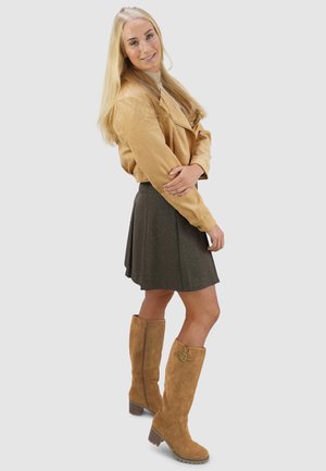 Tan leren cropped jack, lichtbeige top, knielange bruine rok met plooien en tan suede kniehoge laarzen met gespen.