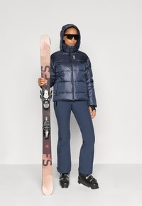 Navy puffer jacket with a hood, navy ski pants, and black ski boots; holding skis with pink accents and bindings. Dark sunglasses worn.