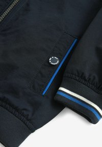 Marinblå jacka med dragkedja, med en blå accentrand och elastiska ärmslut. Inkluderar en rund logoknapp och strukturerad tyg.