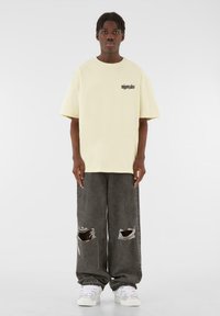 Light yellow oversized t-shirt with black logo on the chest, paired with gray distressed wide-leg jeans and white sneakers.
