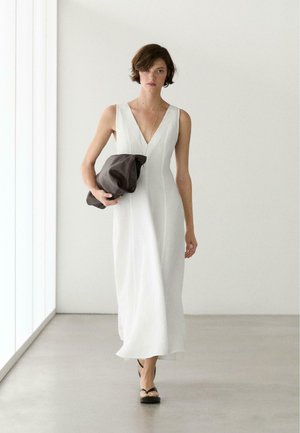 Frau läuft drinnen und trägt ein ärmelloses weißes Maxi-Kleid mit V-Ausschnitt, schwarze Sandalen und hält eine große schwarze Clutch.