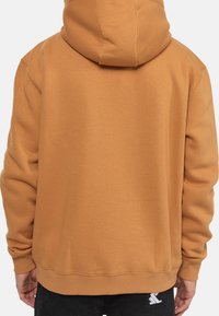 Huvtröja i en varm, brun nyans. Har en avslappnad passform, ribbade mudd och fåll. Synlig sömnad och en liten logotyp på nedre vänstra sidan.