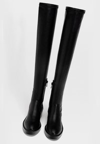 Botas de cuero negro sobre la rodilla con una textura suave, punta redondeada y cremalleras laterales; cuentan con una suela plana y texturizada para mayor tracción.
