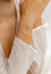 Pulsera de cadena de oro con una pequeña piedra preciosa transparente, llevada en una muñeca. El fondo presenta una funda de tela blanca texturizada.