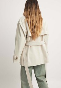 Šviesiai smėlinis paltas su laisvu siluetu, pasižymintis plačiu apykakle, diržu ir sagomis ant rankovių. Dėvimas ant žalių, tekstūruotų kelnėmis.