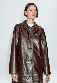 Cappotto in pelle marrone con finitura lucida, caratterizzato da un colletto a punta, chiusura frontale con bottoni e tasche laterali. Maniche lunghe e design minimalista.