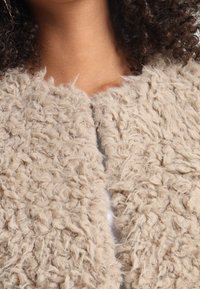 Beige, texturerad fuskpälsplagg med en mjuk, fluffig yta, med en öppen frontdesign och utan synliga knäppningar.