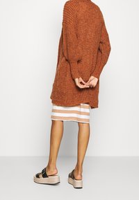 Roodkleurige chunky gebreide cardigan met een relaxte pasvorm, gecombineerd met een gestreepte jurk tot de knie in zacht beige en wit. Zwarte platformsandalen.