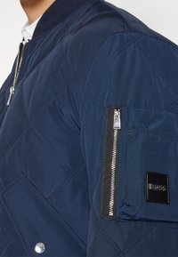 Tmavě modrá prošívaná bomber bunda s kapsou na zip na rukávu, patentními knoflíky a logovou nášivkou; hladká textura a žebrovaný límec.