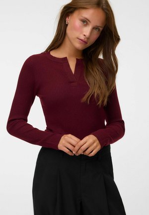 Femme aux longs cheveux bruns portant un pull côtelé bordeaux ajusté avec un col en V et un pantalon noir taille haute, posant les mains jointes.