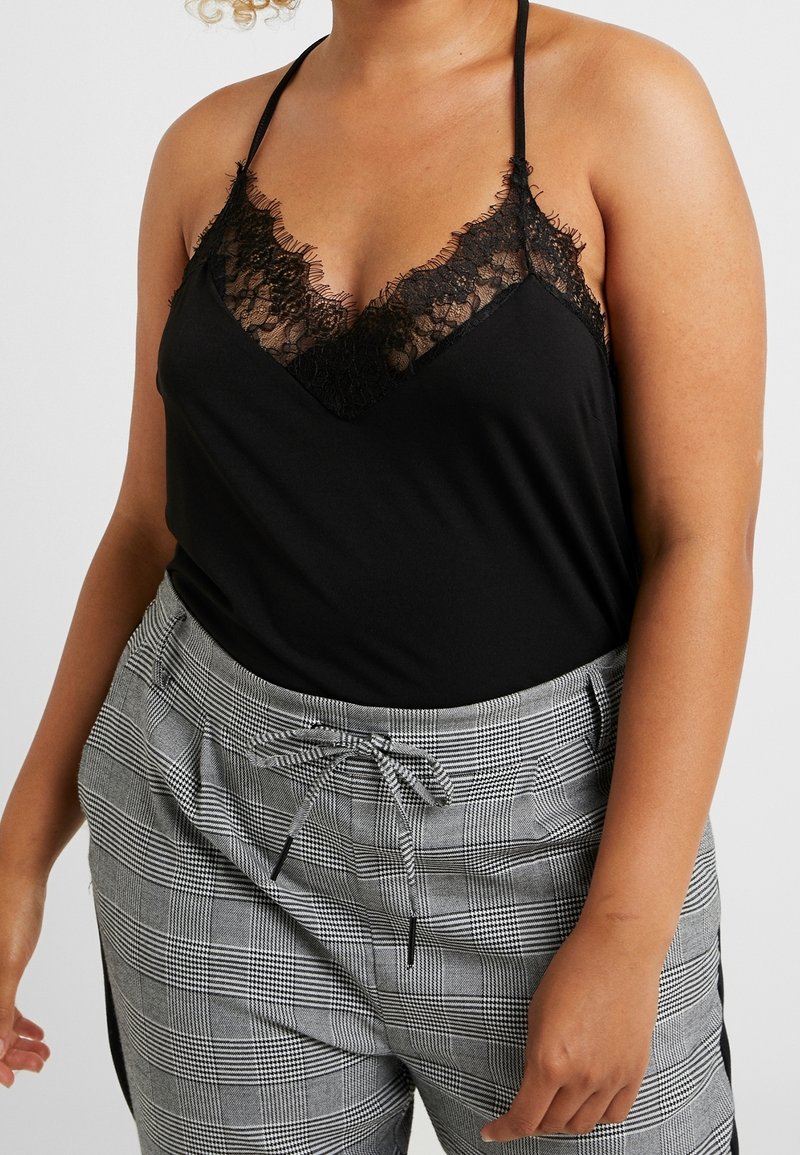Personne portant un caraco noir bordé de dentelle et un pantalon gris à carreaux taille haute avec cordon, vue des épaules aux cuisses.