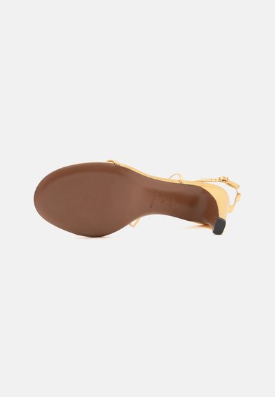 Sandale jaune à talons hauts avec des fines brides, vue de la semelle montrant un dessous marron lisse et un petit talon noir.