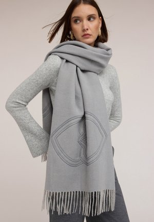 Jonge vrouw met een grote lichtgrijze sjaal met franjes en geometrisch patroon, gedragen over een aansluitende grijze trui en donkere broek, poserend tegen een effen achtergrond.