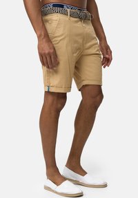 Shorts beiges avec des ourlets roulés, un zip frontal et un accent bleu sur le côté. Associés à des chaussures blanches à enfiler. Ceinture texturée incluse.