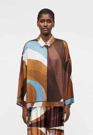 Kobieta ubrana w luźny, blokowy strój z abstrakcyjnymi, zaokrąglonymi wzorami w brązowym, niebieskim, białym i beżowym, stojąca na jednolitym tle.