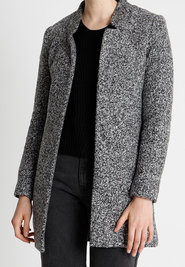 Manteau en laine grise texturée porté sur un pull noir et un pantalon sombre, avec un col montant et des manches longues.