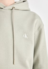 Felpa verde chiaro realizzata in morbido tessuto con tasca frontale. Presenta un cappuccio regolabile con cordino e un piccolo logo bianco sul petto.