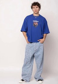 Um jovem está de pé, vestindo uma t-shirt oversized azul com uma estampa de caveira em chamas, calças largas de ganga azul claro com padrão e ténis em preto e branco.