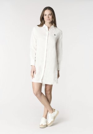 Donna in piedi con le gambe incrociate, indossa un vestito camicia bianco a maniche lunghe e sneakers bianche su uno sfondo bianco uniforme.