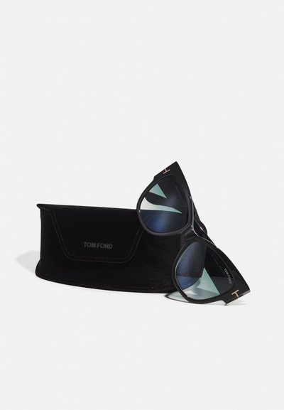 Svarte Tom Ford-solbriller med skrå kattøyeform, blåtonede linser og gullhardware, plassert ved siden av en svart fløyelsdeksel.