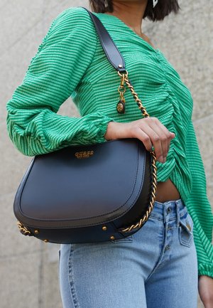Vrouw in lichtblauwe jeans en een groen gestreept shirt, met een zwarte Guess-handtas met gouden kettingdetails over haar schouder.