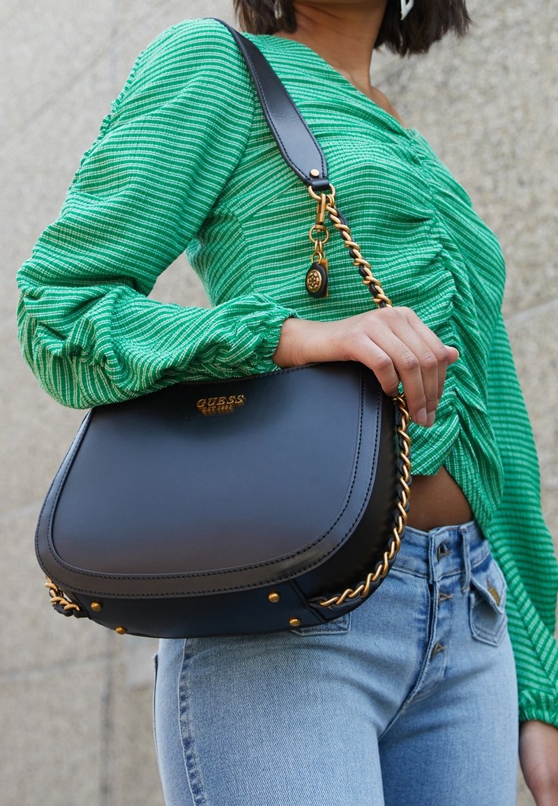 Žena v světle modrých džínách a zeleném pruhovaném topu, držící černou kabelku Guess s zlatým řetízkem přes rameno.