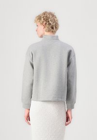 Grå cropped sweatshirt med høy krage, løs passform og mansjetterte ermer, laget av myk stoff, kombinert med et teksturert hvitt skjørt.