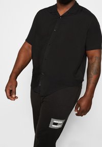 Korte mouwen zwart shirt met een knoopsluiting, gladde stof en een relaxte pasvorm, gedragen met zwarte broek met een witte logodetail.