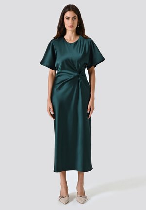 Donna in un abito midi satinado verde scuro con maniche corte, dettaglio a vita torcigliata e tacchi beige a punta su uno sfondo semplice.