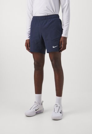Mann trägt marineblaue Sportshorts, weißes Langarmshirt, weiße Kniestrümpfe und weiße Nike-Sneaker mit schwarzem Swoosh-Logo.