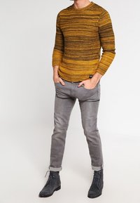 Žlutý a hnědý pruhovaný pletený svetr s dlouhými rukávy, doplněný šedými zúženými kalhotami a černými šněrovacími botami.