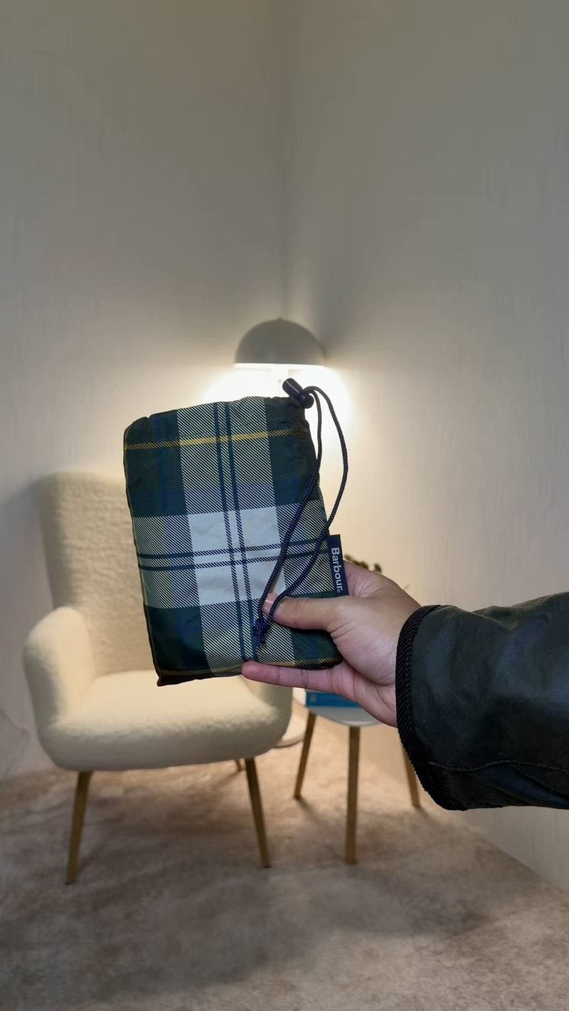 Main tenant une pochette à cordon plissée à carreaux étiquetée Barbour dans une pièce confortable avec un fauteuil blanc et une petite table avec une lampe.