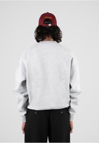 Felpa grigia con vestibilità ampia, polsini a costine e collo a giro. Abbinata a pantaloni neri con due bottoni visibili. Cappellino bordeaux con testo giallo.