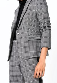 Šedý a černý károvaný blazer s jedním knoflíkem, klopami a ohrnutými rukávy, doplněný odpovídajícími kalhotami.