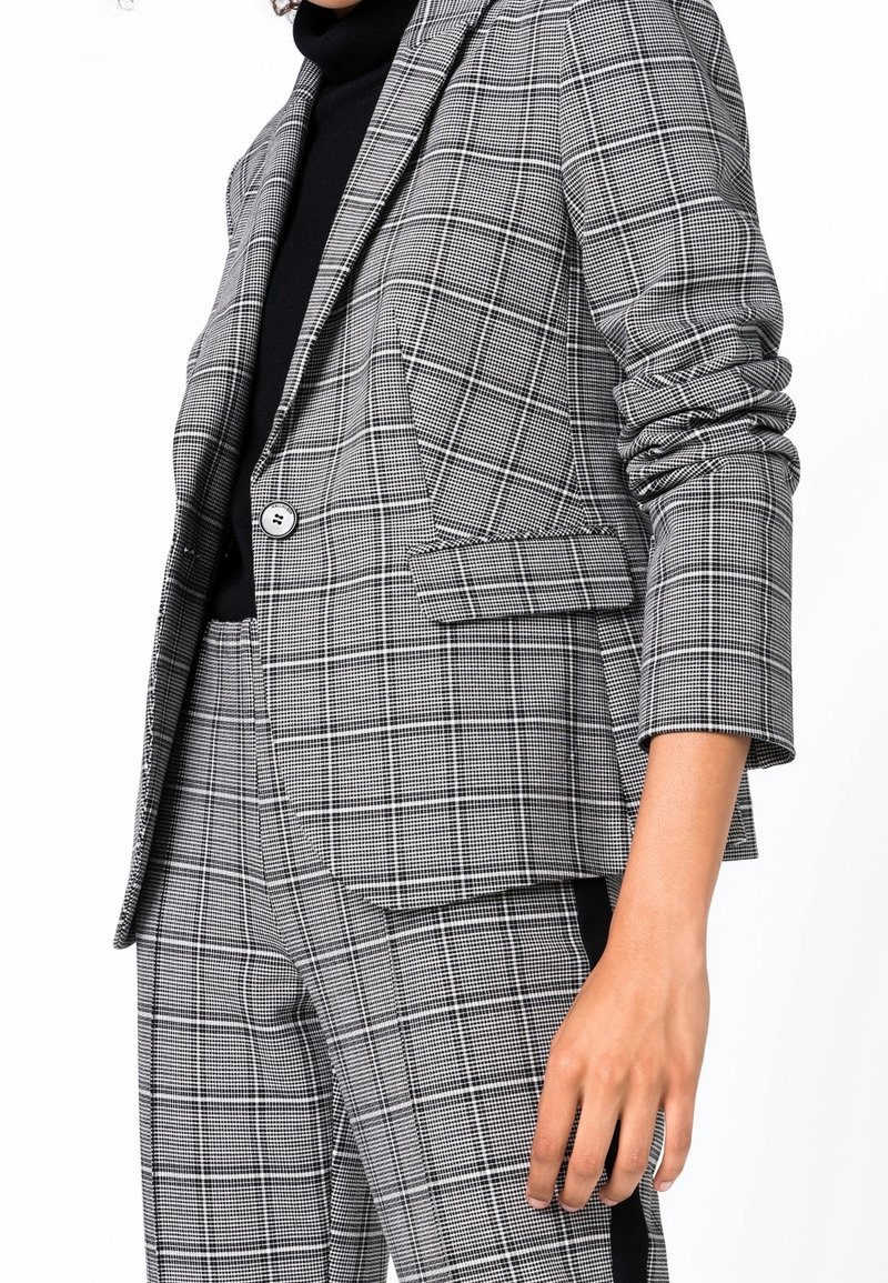 Šedý a černý károvaný blazer s jedním knoflíkem, klopami a ohrnutými rukávy, doplněný odpovídajícími kalhotami.