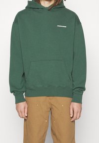 Person klädd i en grön huvtröja med "PEGADOR"-logotyp på bröstet och bruna byxor med sydda fickor, stående mot en enkel vit bakgrund.