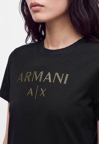 Γυναίκα που φοράει μαύρο T-shirt με χρυσό κείμενο με κουκκίδες που γράφει "ARMANI A|X" στο στήθος, εμφανιζόμενη από τον λαιμό μέχρι τη μέση του κορμού.