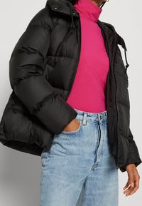 Doudoune noire avec capuche, design matelassé, cordons de serrage attachés, associée à un col roulé rose et un jean taille haute bleu clair.