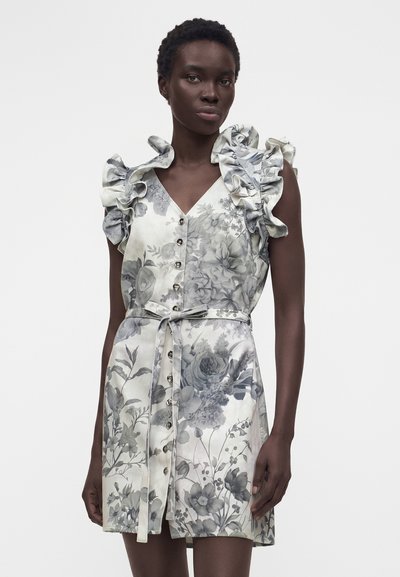 Γυναίκα φοράει αμάνικο φόρεμα με ζώνη και κουμπιά στο μπροστινό μέρος, γκρι λουλουδένιο σχέδιο και βολάν στους ώμους, σε απλό λευκό φόντο.