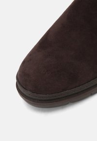 Stivale alla caviglia in pelle scamosciata marrone con punta arrotondata, suola piatta e superficie testurizzata. Presenta un dettaglio di cuciture sottili lungo la suola.