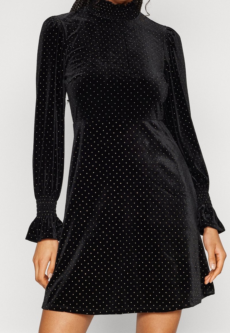 Robe en velours noir avec de longues manches bouffantes, des poignets à volants, un col haut smocké et de petits pois dorés espacés de manière régulière.