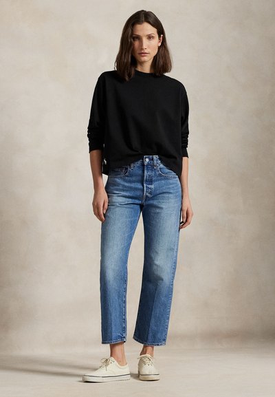 Nő fekete hosszú ujjú pulóvert, magas derekú kék farmert és fehér tornacipőt visel, semleges háttér előtt áll.