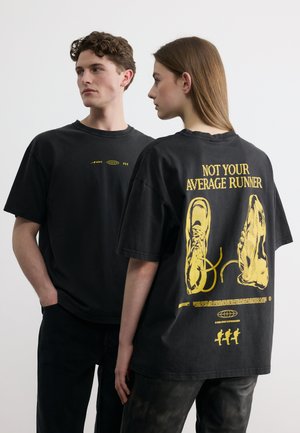 Junger Mann und Frau tragen schwarze, übergroße T-Shirts; auf dem Rücken der Frau sind gelbe Laufschuhe und der Schriftzug „NOT YOUR AVERAGE RUNNER“ zu sehen.