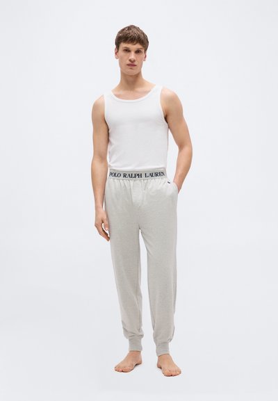 Νέος άντρας όρθιος ξυπόλητος, φορώντας λευκή αμάνικη φανέλα και ανοιχτό γκρι παντελόνι φόρμας με την ελαστική ζώνη της Polo Ralph Lauren.