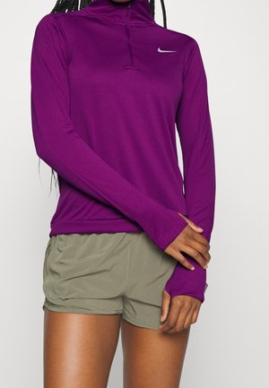 Personne portant un haut Nike violet à manches longues avec des trous pour les pouces et un short de sport vert olive, debout, les bras croisés à la taille.