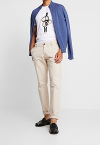 Lekka niebieska kurtka zapinana na zamek, na białej koszulce z czarno-szarym nadrukiem. Do tego beżowe chinosy i czarne mokasyny, które dopełniają stylizację.