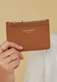 Tan leather cardholder with a textured surface, gold embossed logo, zippered compartment, and multiple card slots on the front.